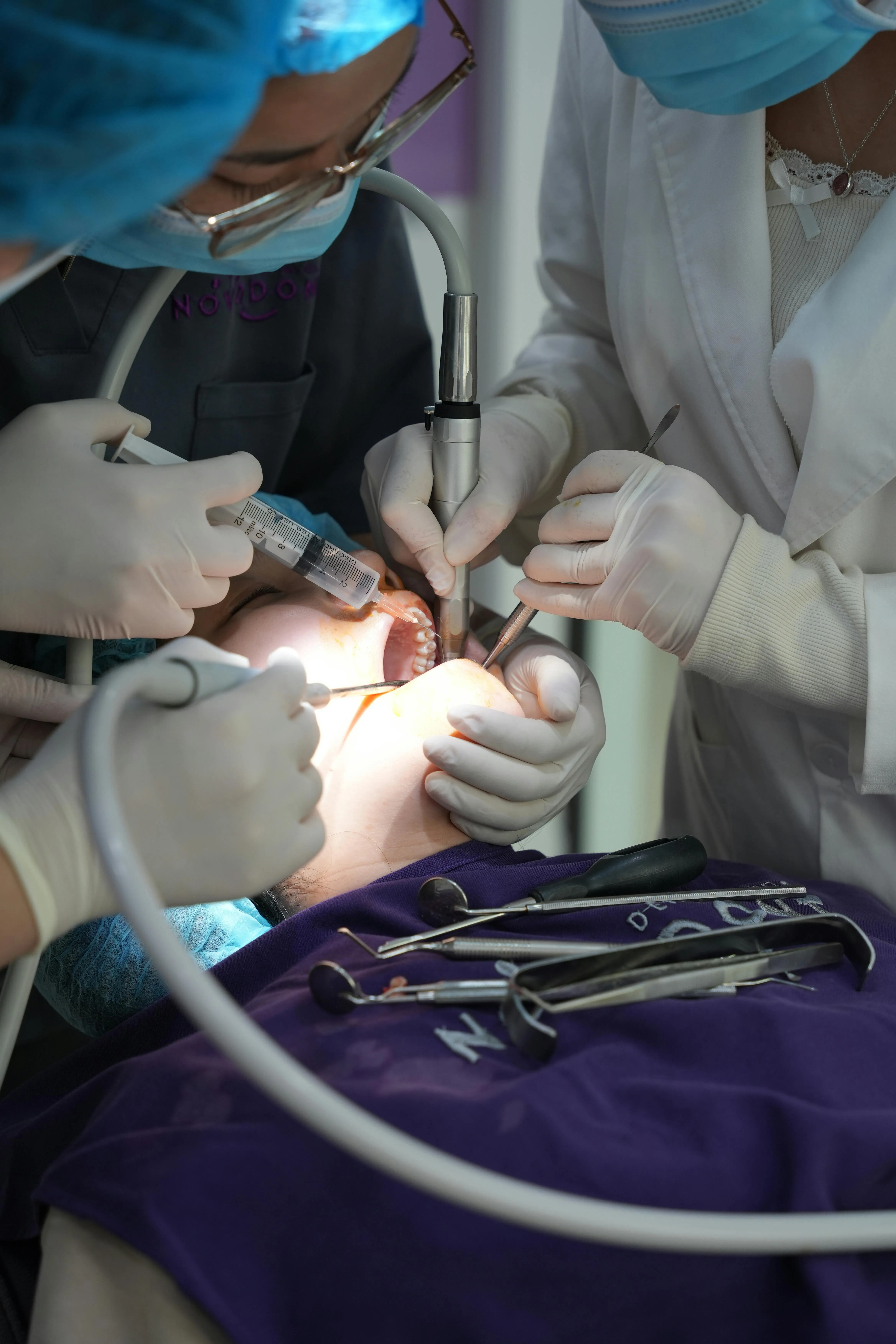 Dentists wearing gloves and masks treat a patient's teeth using dental instruments and a syringe.
