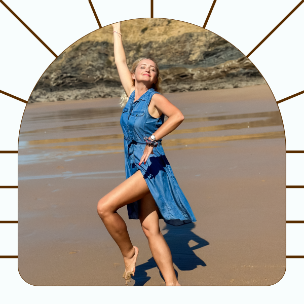Woman in a sleeveless blue dress posing barefoot on a sandy beach with rocky cliffs in the background, holding sunglasses up in her right hand.