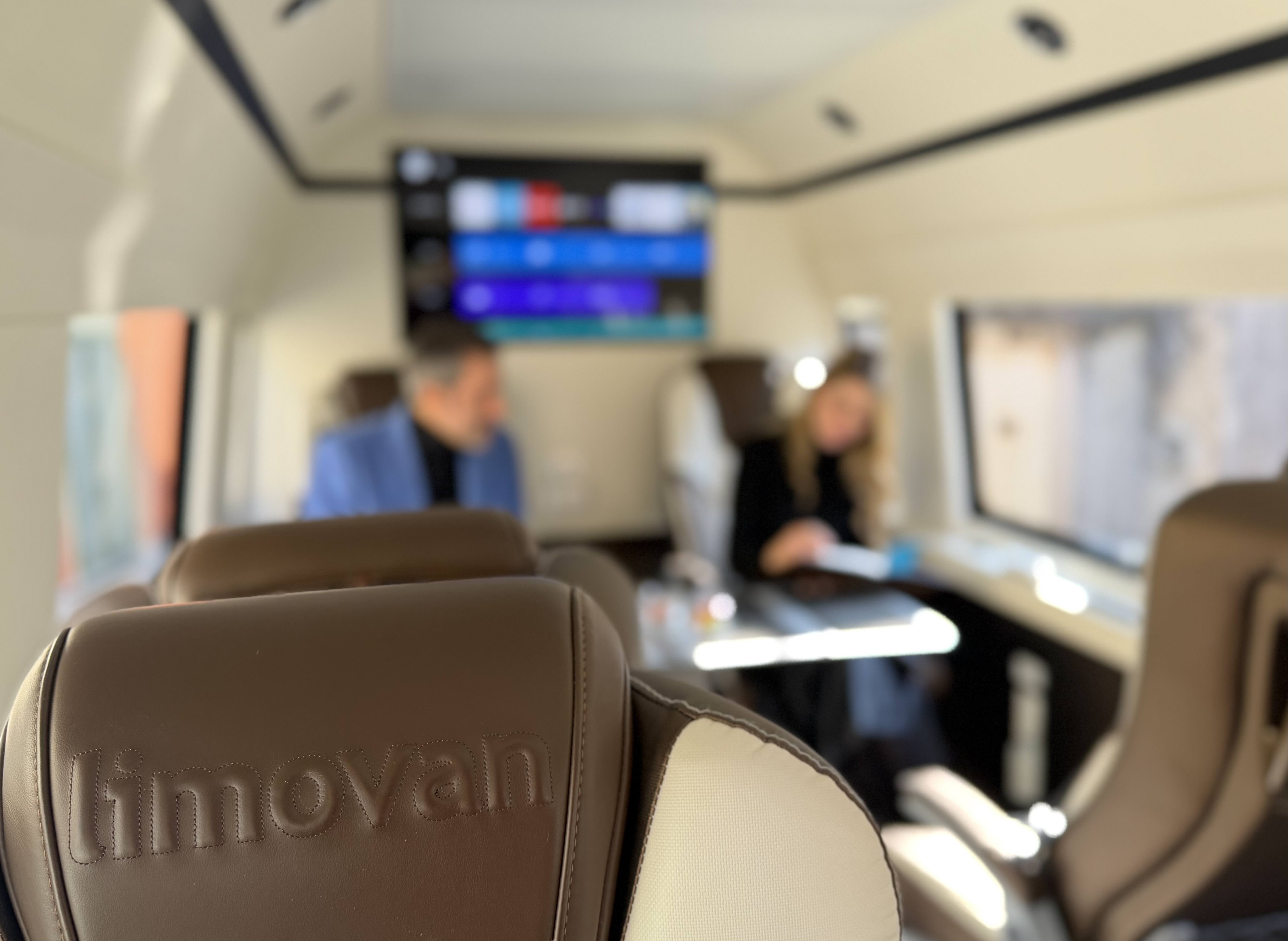 Branded seat view inside the Moveolux Limovan Light Edition with passengers in the background