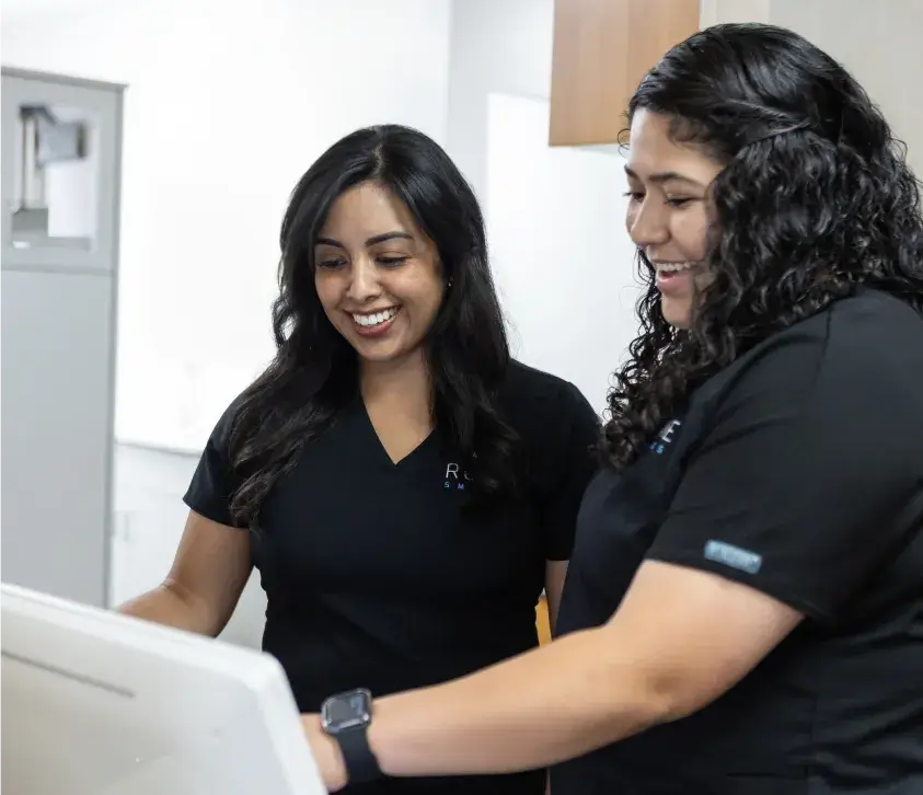 Two women in matching black shirts smiling and looking at a computer monitor.