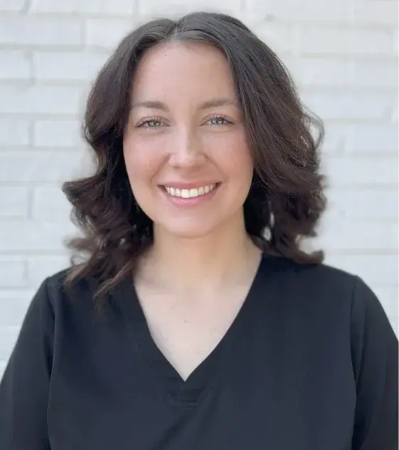 Smiling woman with shoulder-length brown hair, wearing a black shirt, stands against a light brick wall.