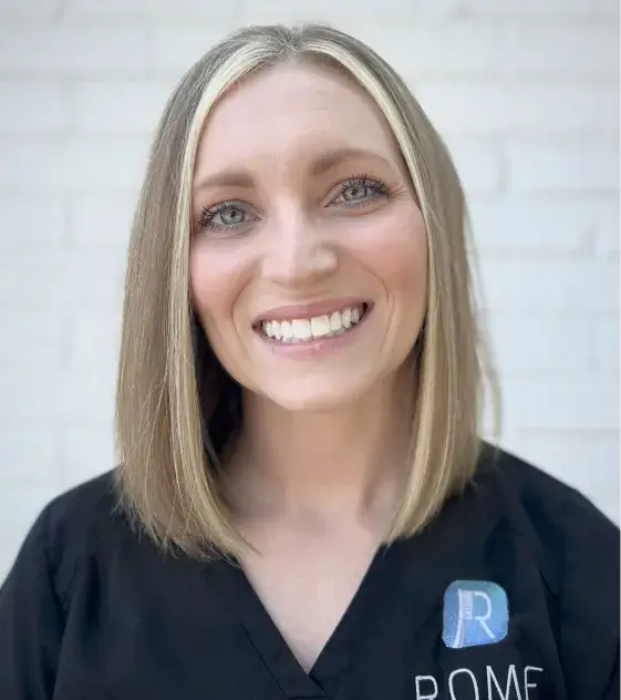 Person smiling, wearing a black shirt with a logo and standing in front of a plain background.