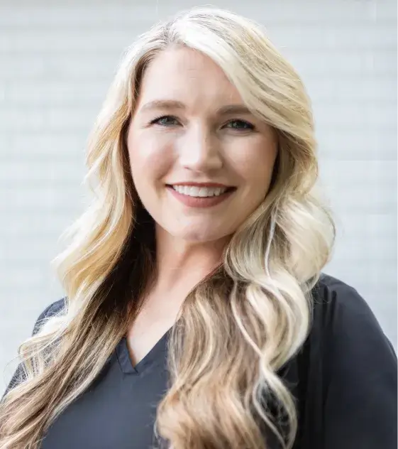 Smiling woman with long blonde hair wearing a black shirt stands in front of a light background.