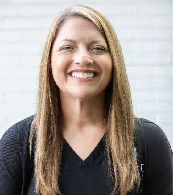 A woman with long hair smiles directly at the camera against a plain background.