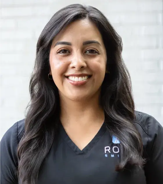 Smiling person wearing a black top with embroidered logo stands in front of a light-colored wall.