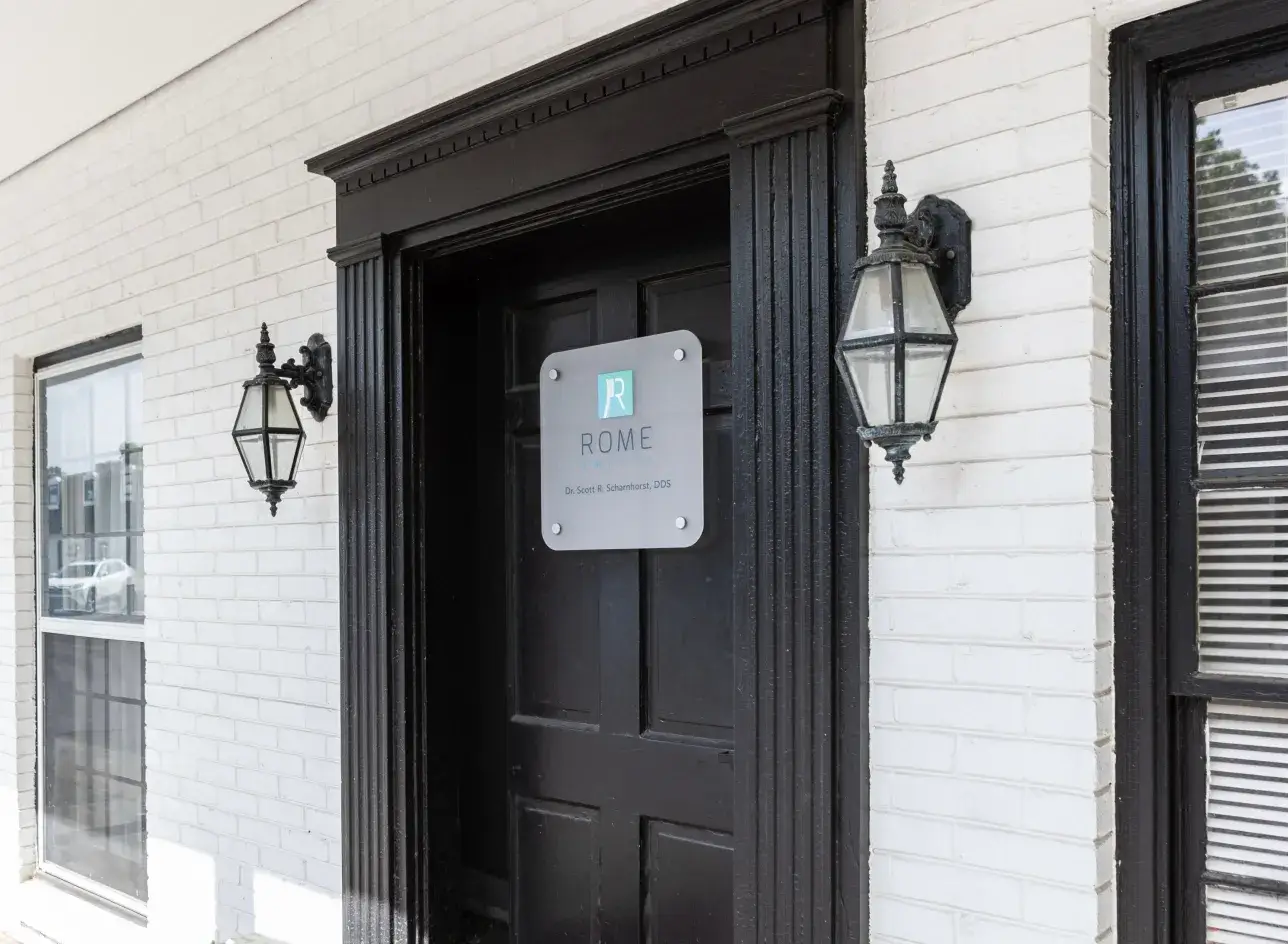 A black door with a sign labeled "Rome" is flanked by two lanterns on a white brick wall.