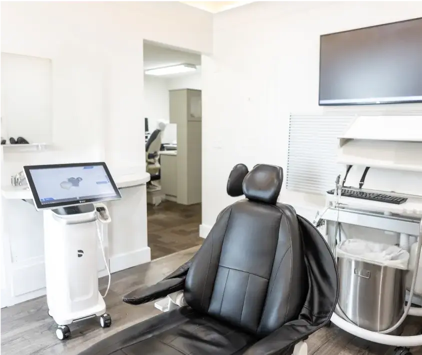 A black dental chair is in a modern dental office with screens and equipment nearby.