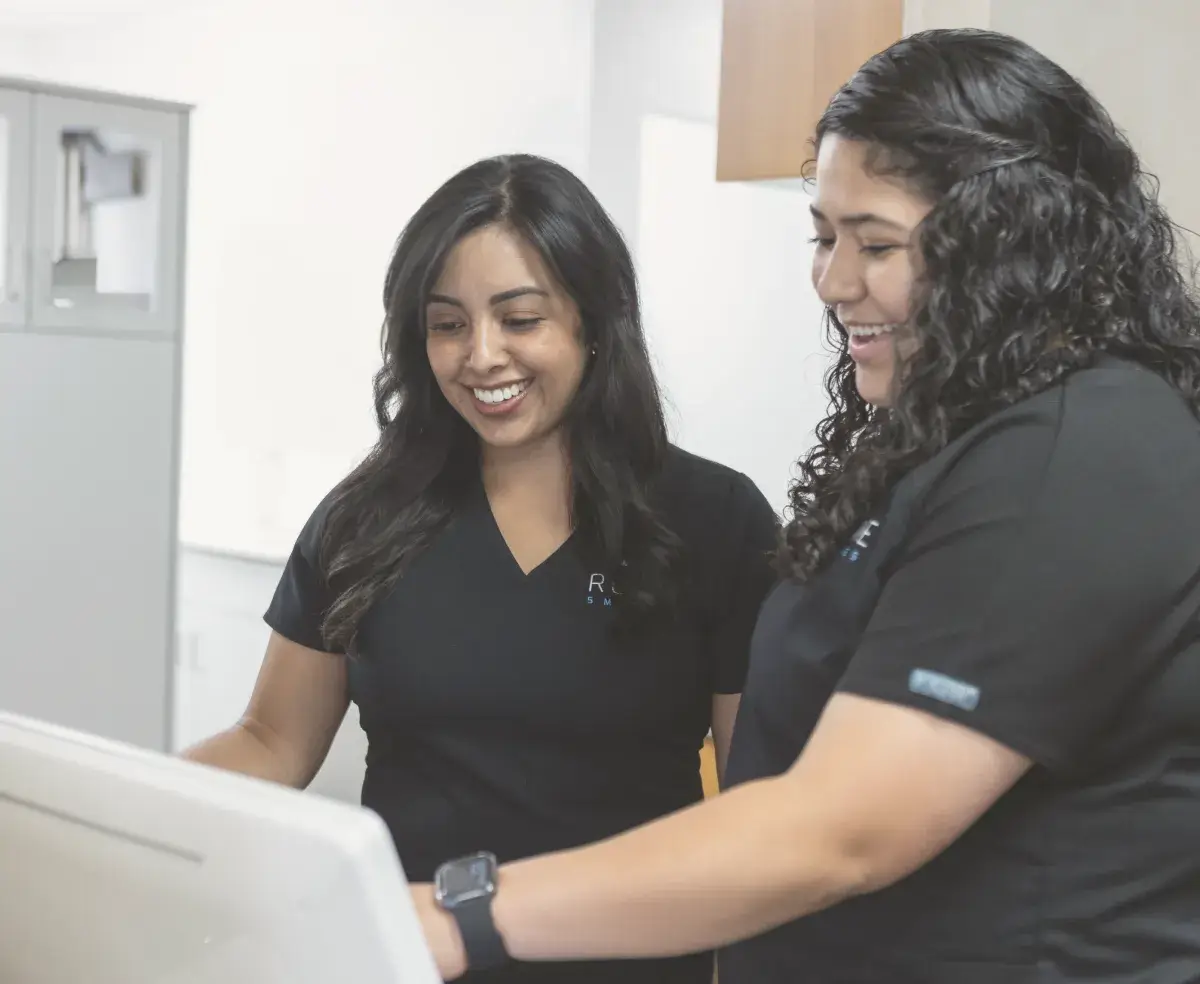 Two women wearing black uniforms smile while looking at a computer screen in an office setting.