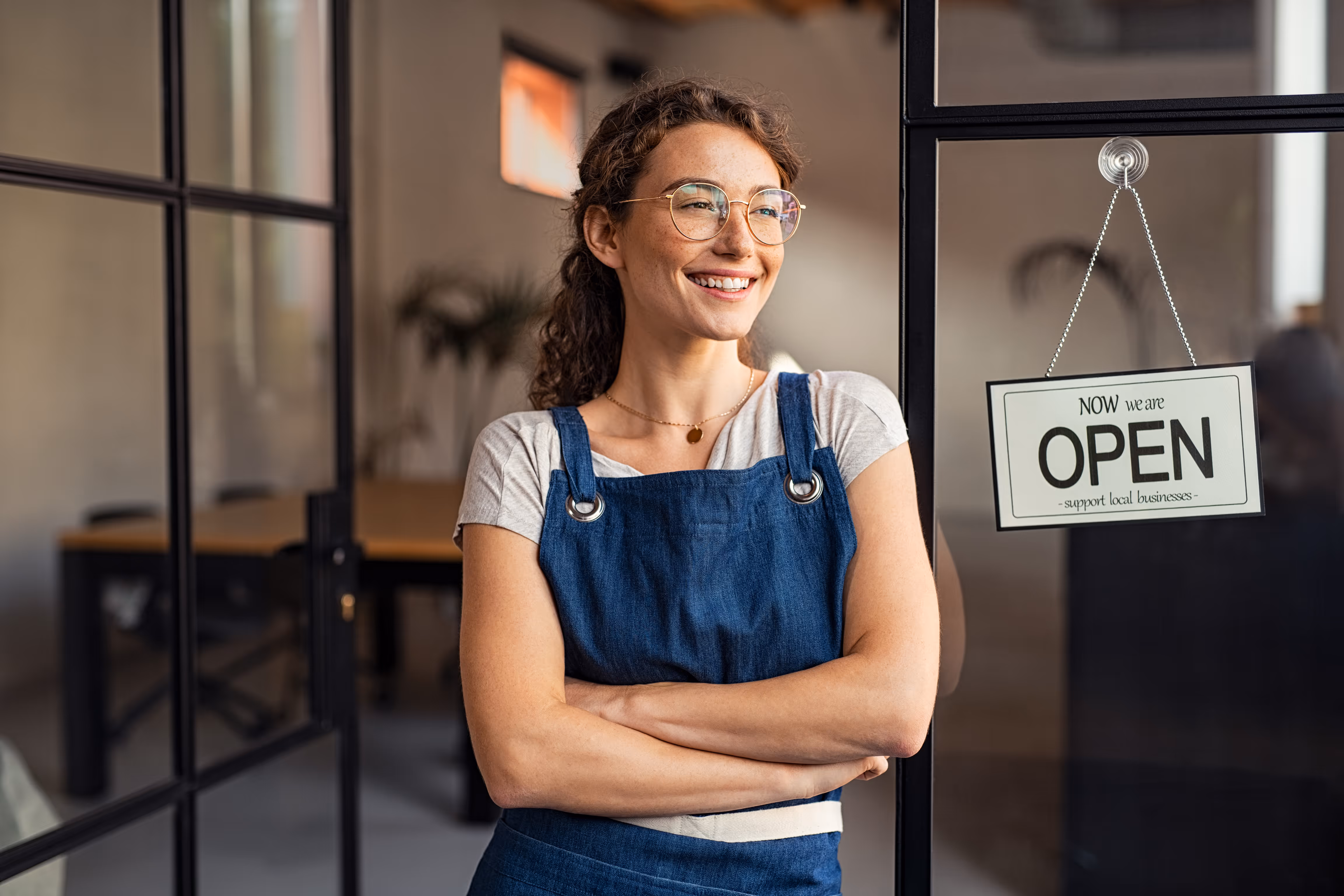 A smiling woman with glasses and an apron standing next to a sign that says "We are now open"