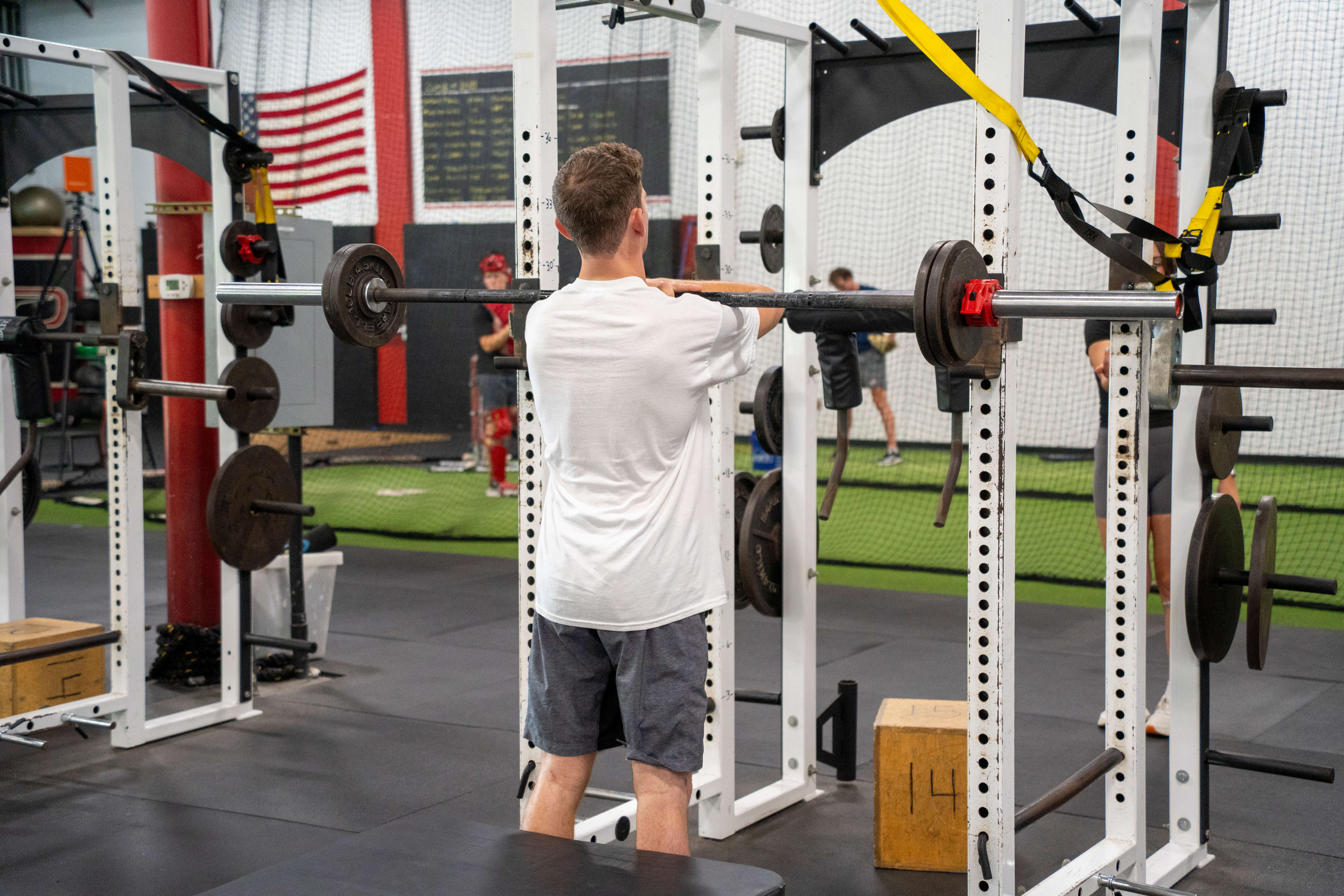 Man in white shirt and gray shorts lifts a barbell in a gym with an American flag in the background.
