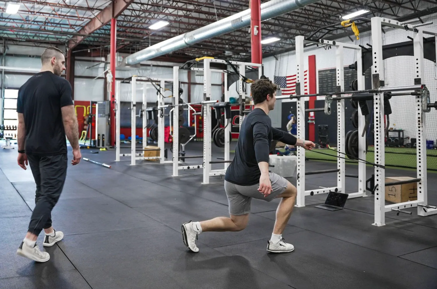 A man in gray shorts and black top doing resistance band exercises in a gym while another man in black clothes watches.
