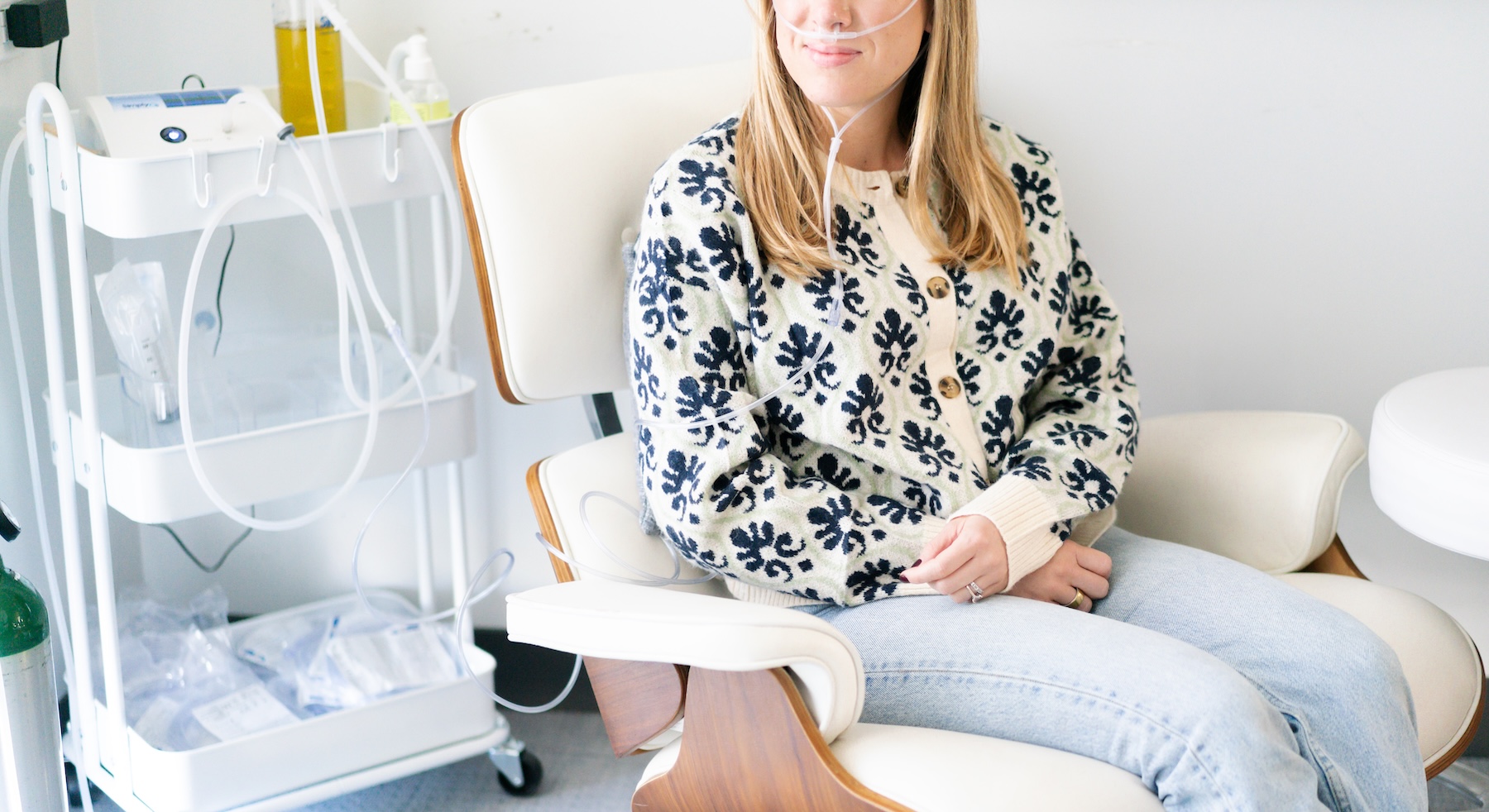 A woman wearing a patterned sweater and light jeans sits in a white cushioned chair with wooden armrests, receiving ozone insufflation through clear nasal tubing connected to a medical device on a nearby cart.