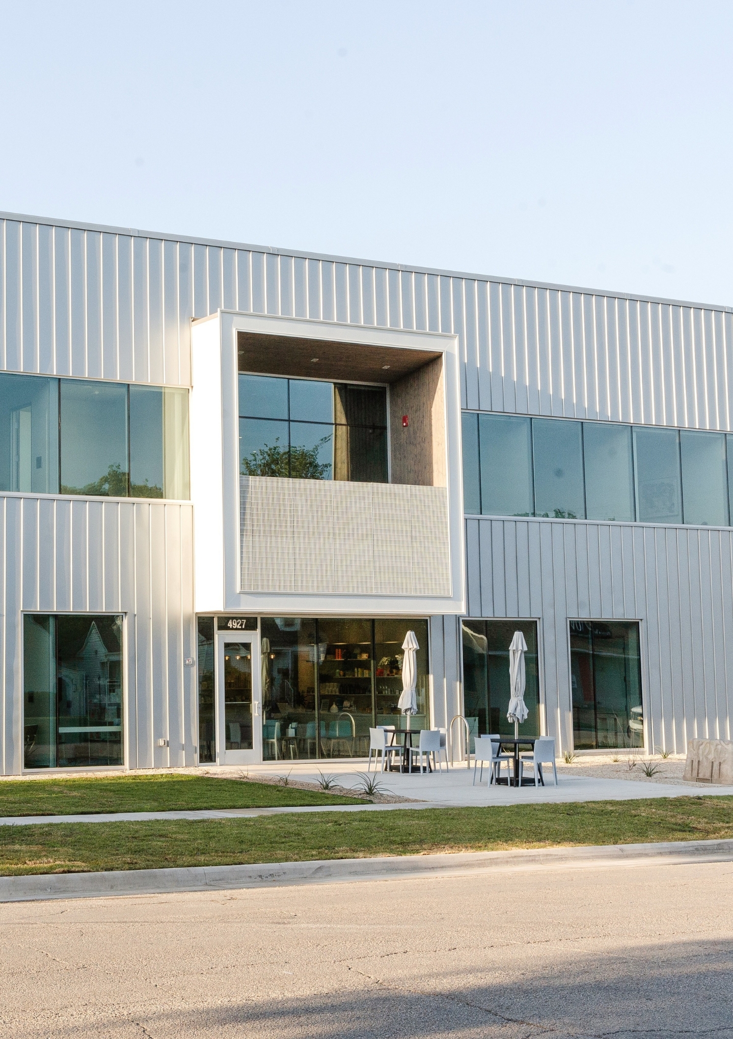 Modern two-story building with a metallic facade, large glass windows, and an inset balcony above the entrance with the address number 4927. Outdoor seating area with tables, chairs, and closed umbrellas on a concrete patio in front of the building. Grass lawn and a paved street are visible in the foreground.