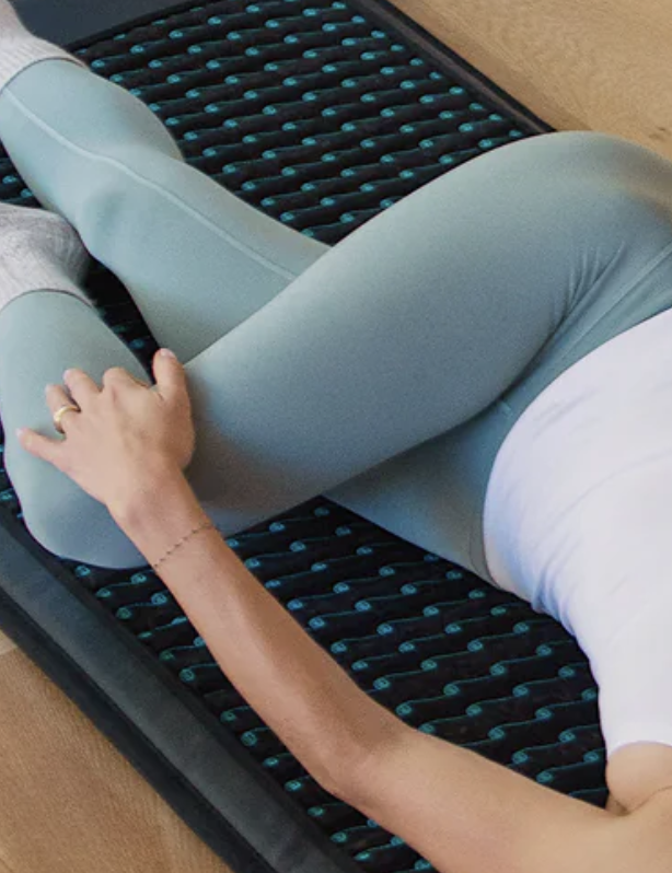 Person lying on their side on a textured black and blue exercise mat, stretching their bent leg while wearing light green leggings and a white top.