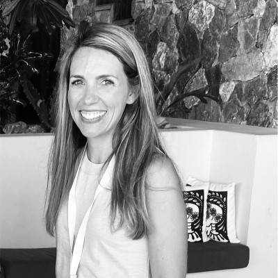 A person stands smiling, wearing a light sleeveless top and lanyard. They are indoors, in front of a textured stone wall with decorative cushions nearby.