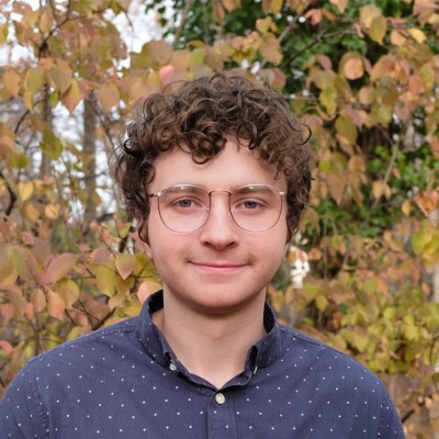 A person with curly hair and glasses looks directly at the camera, standing outdoors. The background features trees with autumn-colored leaves.