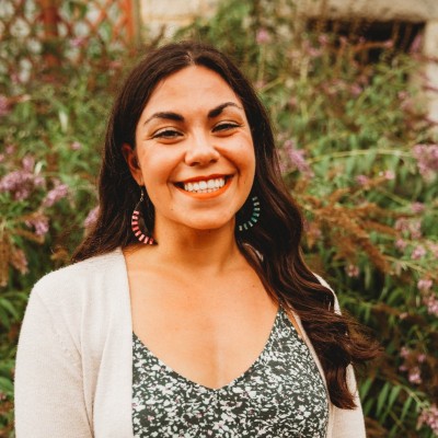 A person smiles broadly, standing in front of lush greenery with pink flowers. They wear a patterned dress, white cardigan, and vibrant earrings, conveying a cheerful and warm atmosphere.