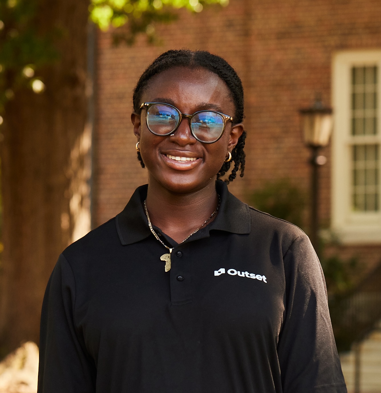 Person smiling, wearing glasses and a black polo shirt with "Outset" logo, stands outdoors. Background includes a tree, brick wall, and window. The setting is daylight with natural light.