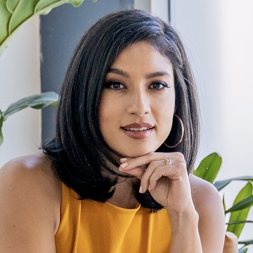 A person wearing a yellow top rests their chin on their hand, seated near a window with green plants in the background.