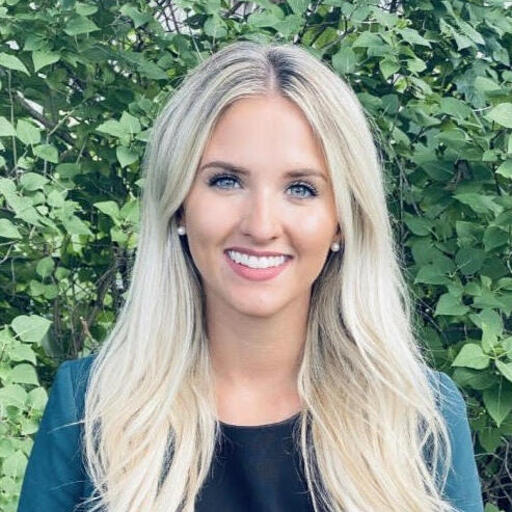 A person with long, blonde hair smiles while standing against a background of lush green foliage. 