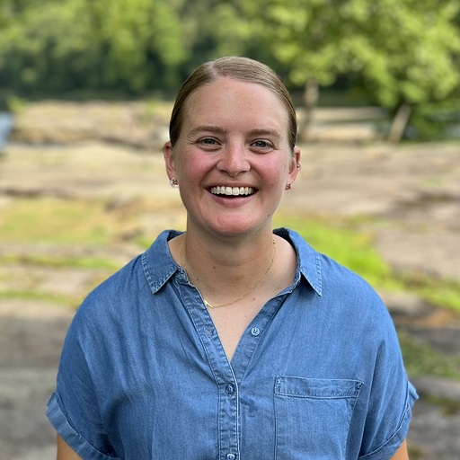 A person is smiling while standing outdoors, wearing a blue button-up shirt. The background features blurred greenery and rocks, suggesting a natural, park-like setting.