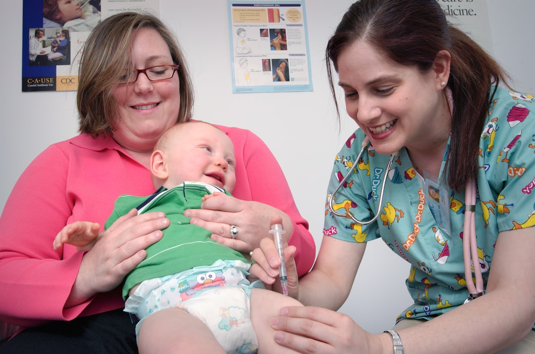 A nurse administers a vaccine to a smiling baby held by a woman in a medical office, with colorful posters on the wall in the background.