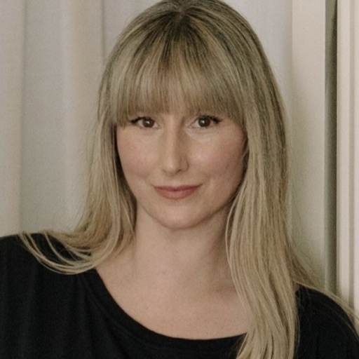A person with long, light-colored hair and bangs is looking directly at the camera, smiling slightly. They are wearing a black top, standing in front of a neutral curtain background.