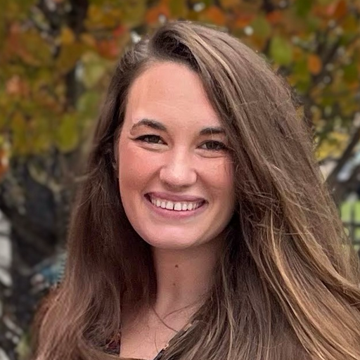 A person smiles, standing in front of a tree with autumn leaves.