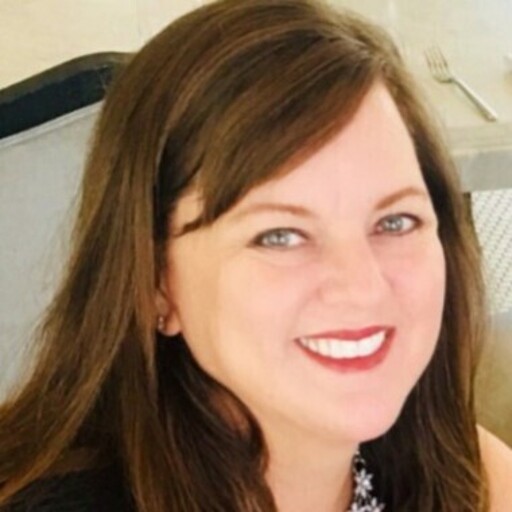 A person with long brown hair is smiling while sitting in a light-colored room. There's a fork visible on the table behind them.