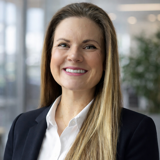 A person is smiling while looking at the camera, wearing a dark blazer and white shirt. The background shows an office setting with a blurred plant and modern decor.