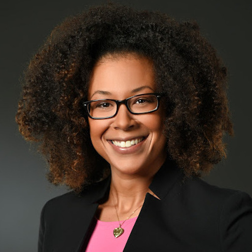 A person with curly hair and glasses smiles confidently, wearing a pink top and a dark blazer, set against a plain dark background.