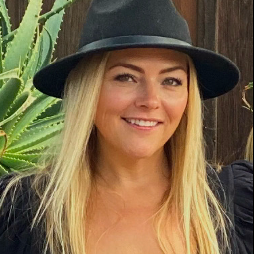 A person wearing a wide-brimmed black hat and black shirt is smiling outdoors, with green aloe plants and a wooden fence in the background.