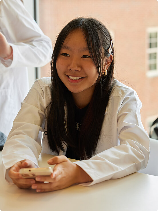 A group of future medical students wearing white lab coats gather around a table, smiling and using smartphones. Nearby are black bags labeled "Class of 2026" in a bright room with large windows.