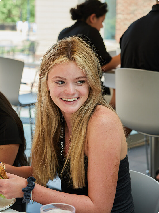 A future medical student with long blond hair is smiling while sitting at a table, holding food. The context is a casual indoor dining area with other people in the background.