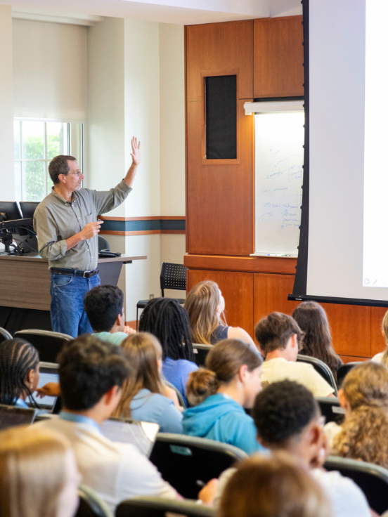 Instructor gestures while presenting to students in a lecture hall. A projection screen displays information, with attentive students seated and taking notes. Classroom setting with natural light streaming in.
