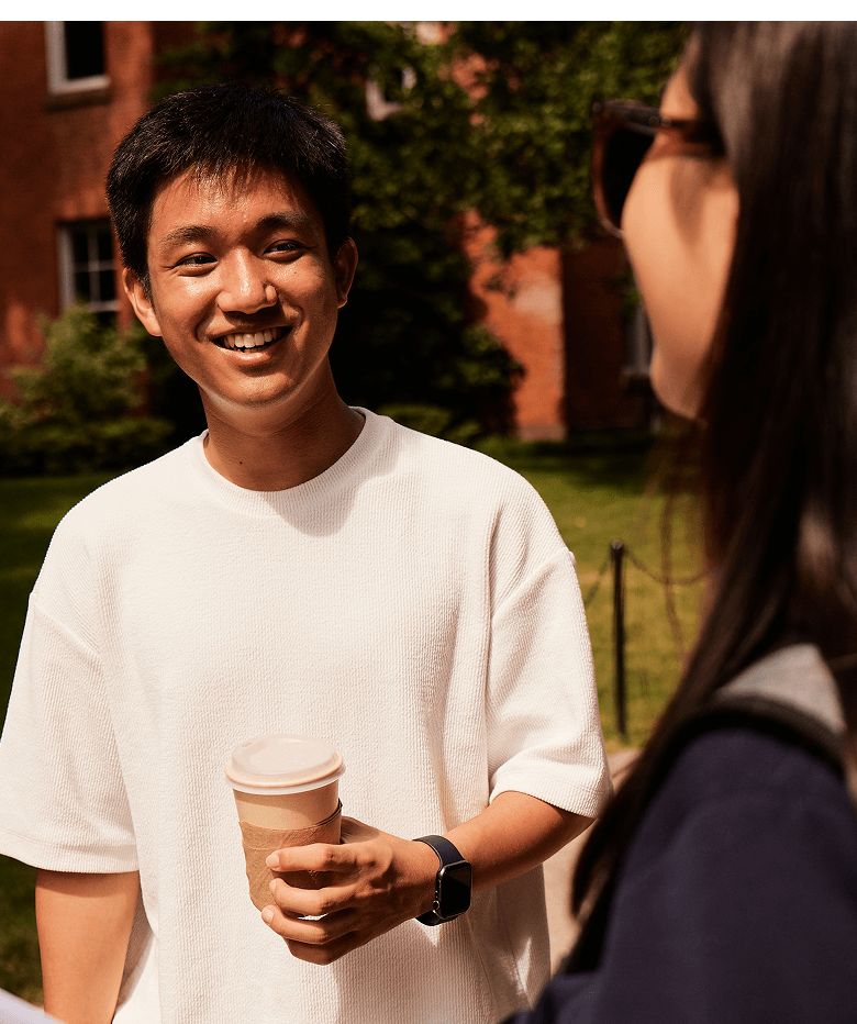 A student interested in medicine holds a coffee cup and smiles while talking to another student outdoors, with a brick building and greenery in the background.