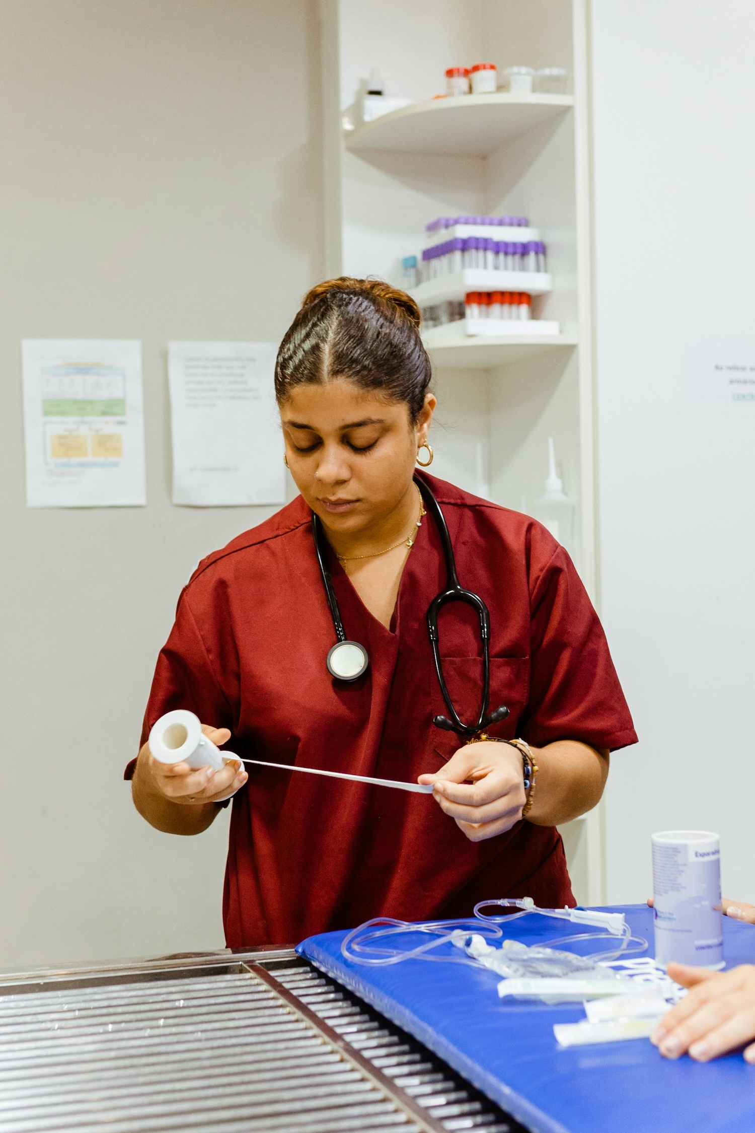 A doctor in a maroon scrub with a stethoscope around their neck is pulling medical tape. They stand at a table with medical supplies, surrounded by shelves and paperwork.