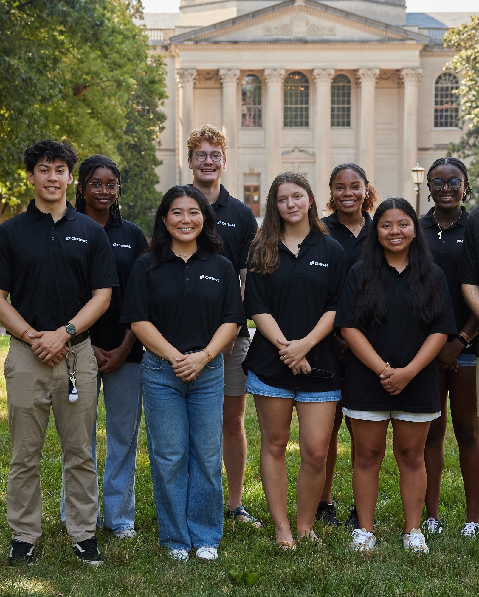 A group of eight Outset staff members stands smiling, wearing black shirts with "Outset" logos. They are outdoors on grass in front of a grand, columned building.