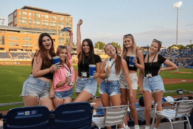 Six people holding beverages stand and smile in a stadium overlooking the baseball field, with empty chairs in front and a building in the background under a clear sky.
