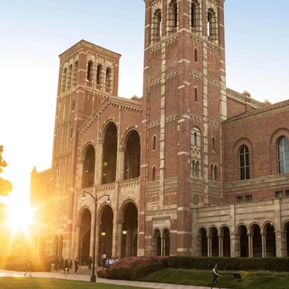 A grand, red-brick building with arched windows and towers stands majestically. People walk in front as the sun sets, creating a warm glow in the surrounding campus environment.