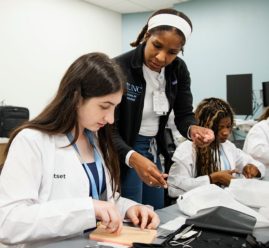 A student practices suturing on a model while an instructor guides her in a classroom setting. The student wears a lab coat, and the instructor's coat reads "UNC School of Medicine."