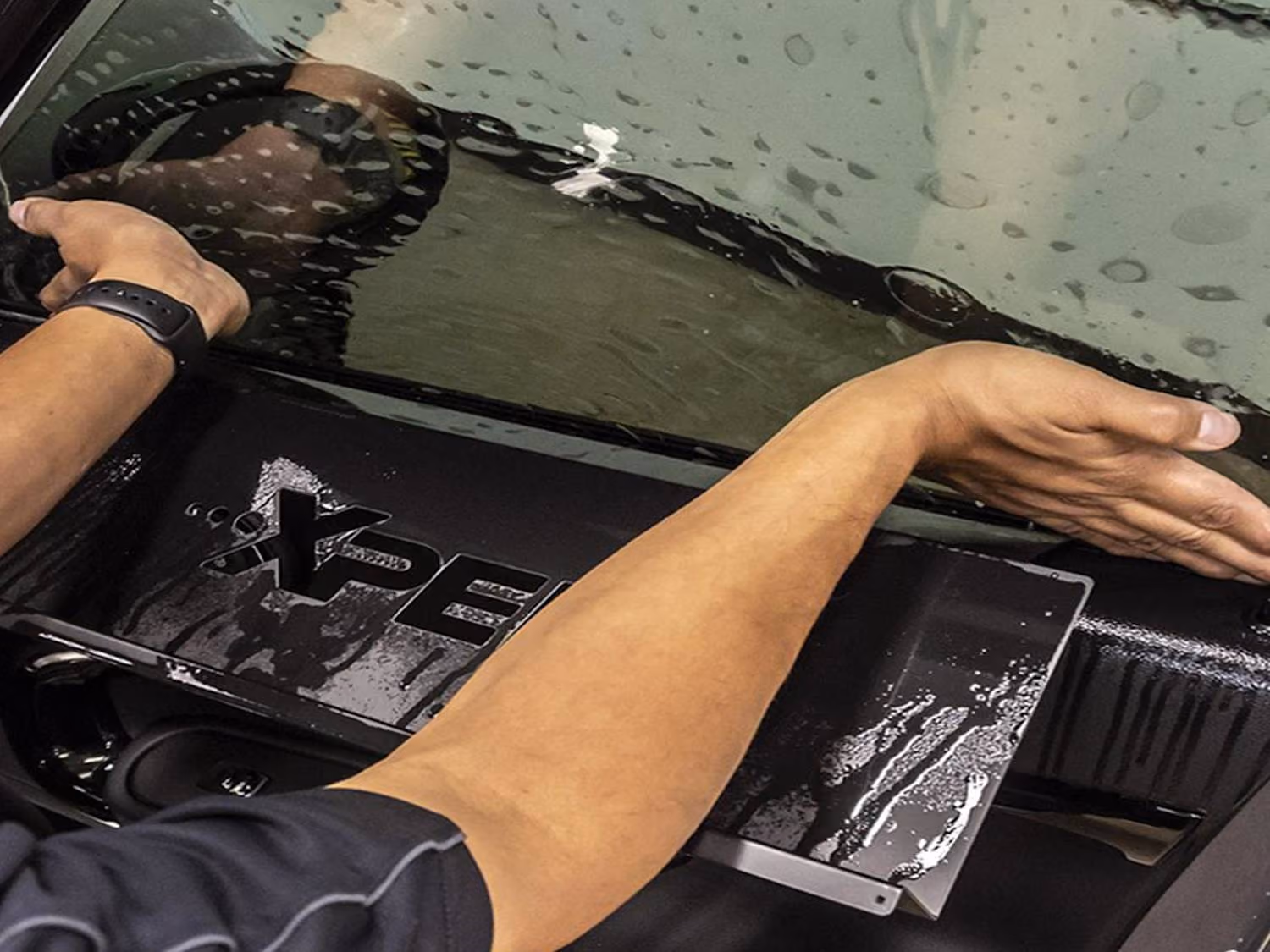Two hands smoothing a wet black XPEL protective film on a car window surface.