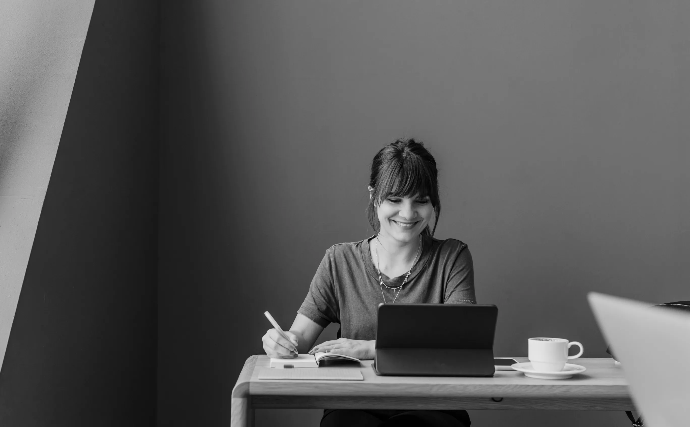 Smiling woman with bangs sitting at a desk, writing in a notebook and looking at a tablet, with a cup of coffee on the table.
