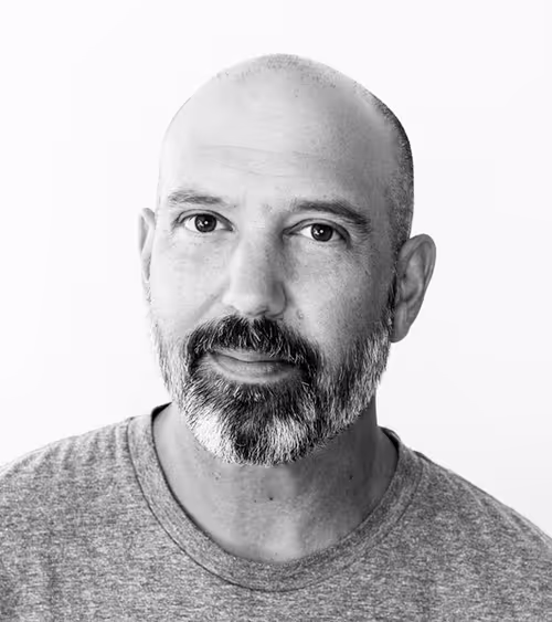 Black-and-white portrait of a middle-aged bald man with a full beard wearing a gray t-shirt, looking directly at the camera.