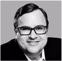 Black and white portrait of a smiling man wearing rectangular glasses and a suit jacket.