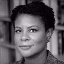 Headshot of a woman with short curly hair wearing earrings, smiling gently in front of blurred bookshelves.