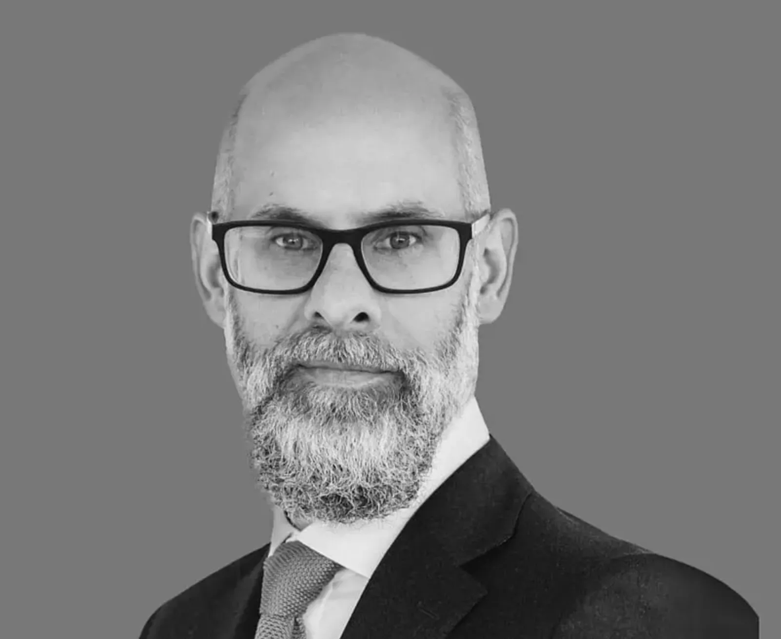 Portrait of a bald man with a gray beard, wearing black-rimmed glasses, a suit jacket, white shirt, and tie against a plain gray background.