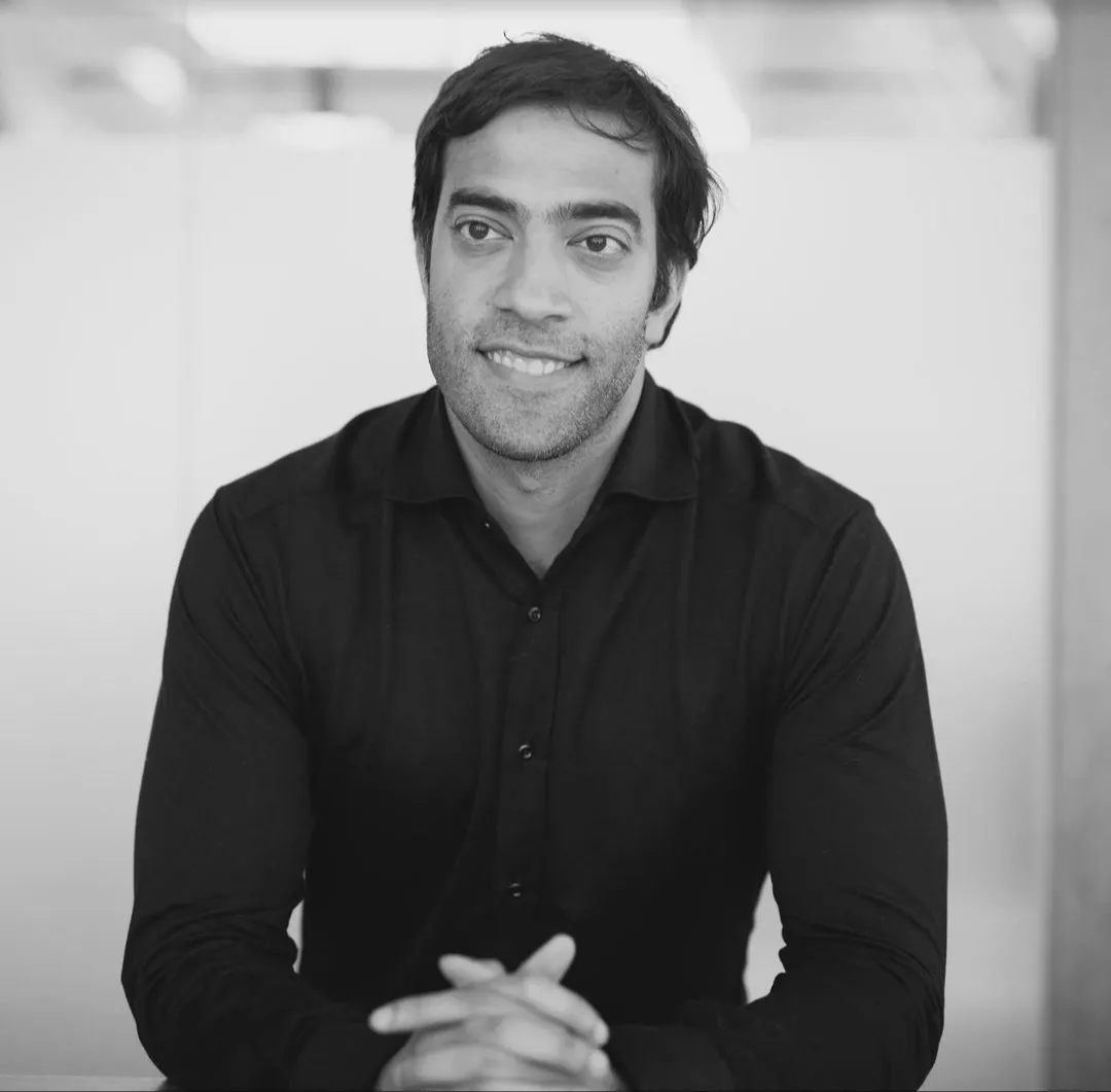 Smiling man with short dark hair wearing a black button-up shirt sitting with hands clasped.