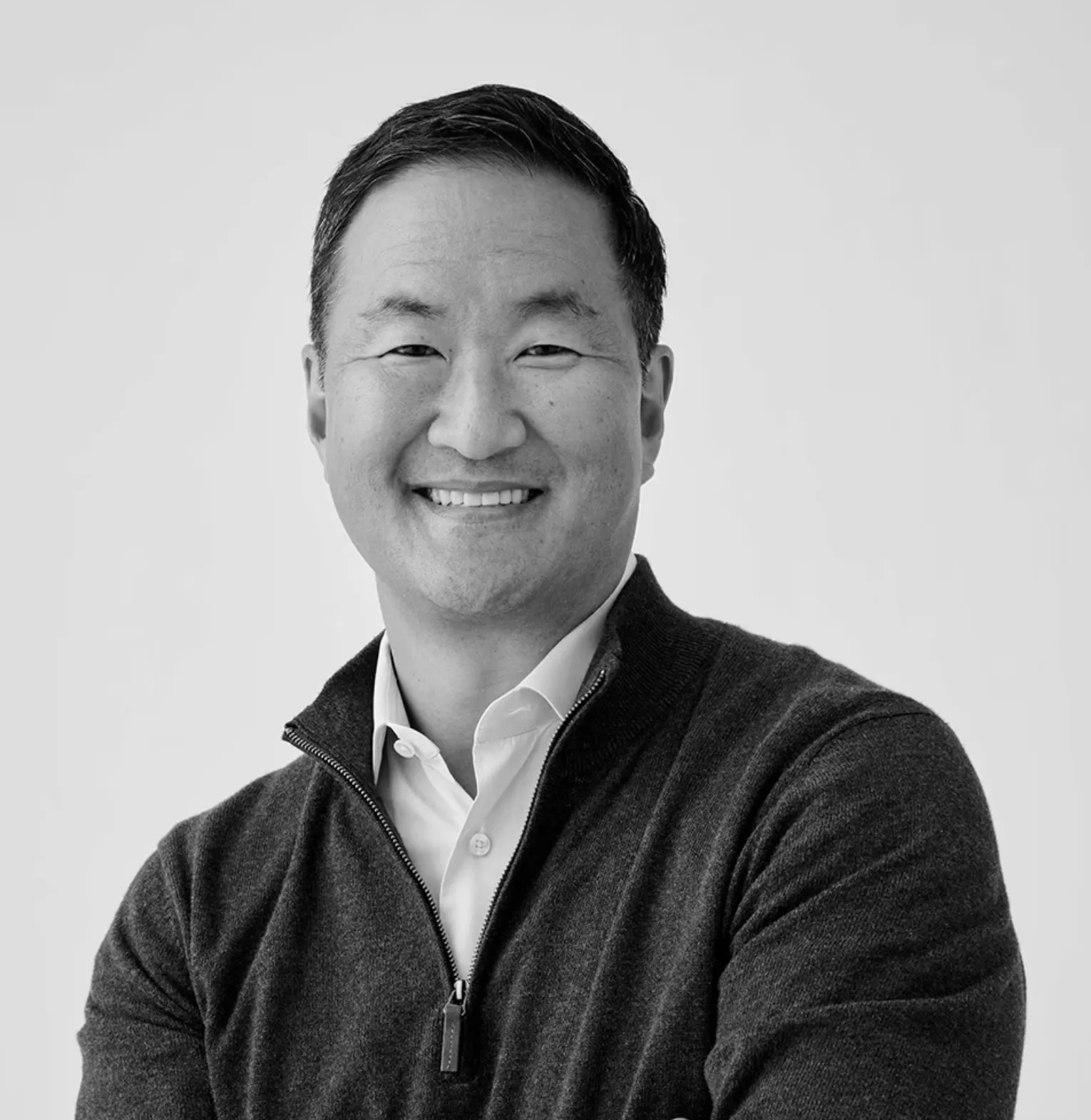 Smiling man with folded arms wearing a dark zip-up sweater over a white collared shirt.
