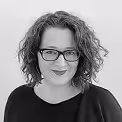 Smiling person with curly hair wearing glasses and a dark top against a white background.